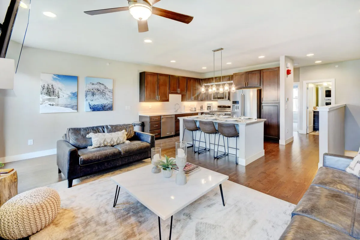 A warm and inviting living space with a plush sofa, modern coffee table, and a glimpse of the kitchen island. Artwork adorns the wall, and the overall feel is perfect for family gatherings or relaxing evenings. The space feels both luxurious and comfortable, echoing the mountain-lodge vibe.