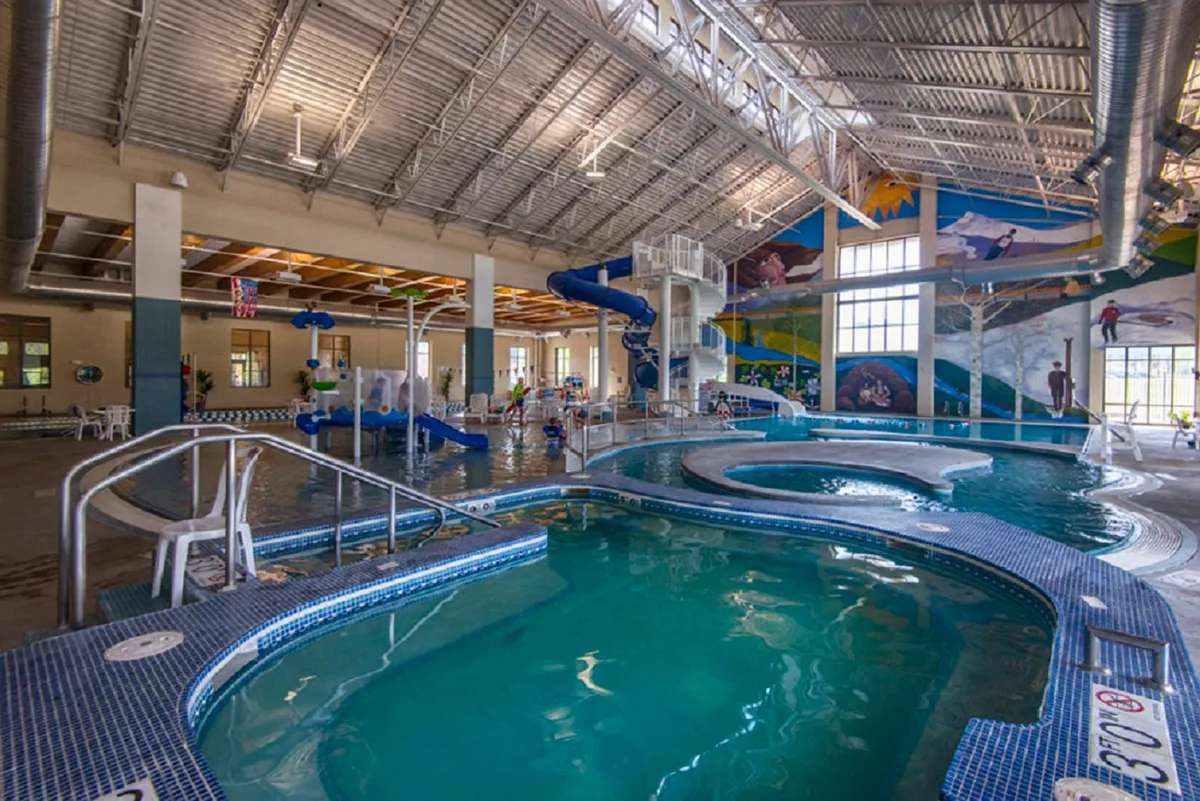The vibrant indoor pool area at the Grand Park Rec Center, featuring a colorful waterslide, a swimming pool, playful murals, and children laughing and playing in the water. The energy is joyful and inviting, perfect for families.