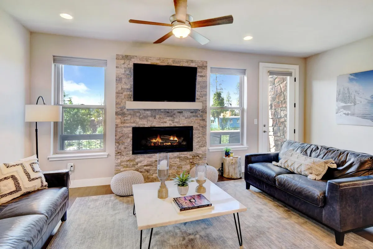 The image showcases a warm and inviting living room featuring a crackling fireplace, plush couches arranged for conversation, a sleek TV, and large windows that fill the space with sunlight and offer scenic views. The room radiates comfort, perfect for cozy winter evenings or sunlit relaxation with family or friends.