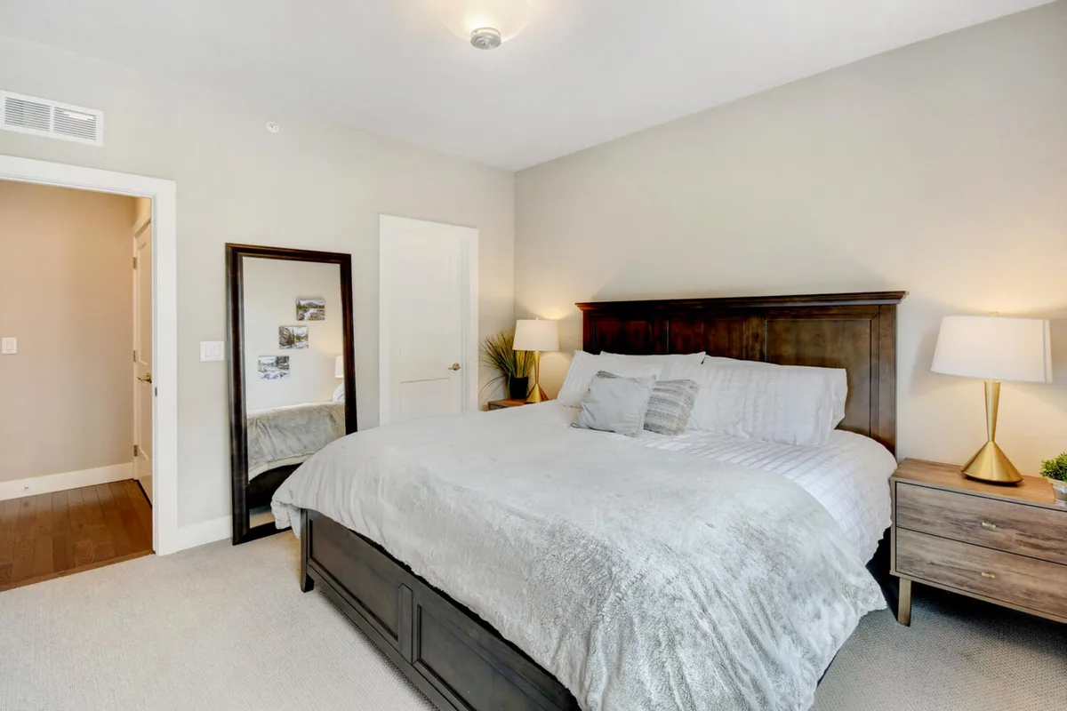 This image shows a serene bedroom with a plush bed, a nightstand with a lamp, and a large mirror, all softly lit by natural light streaming through the window. The atmosphere is calm and inviting, emphasizing comfort and relaxation.