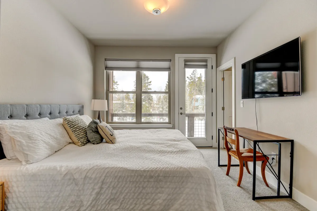 A comfortable king-size bed is set beneath large windows that frame beautiful outdoor views. The room includes a small desk with a chair and a television, offering versatility for both relaxation and entertainment.