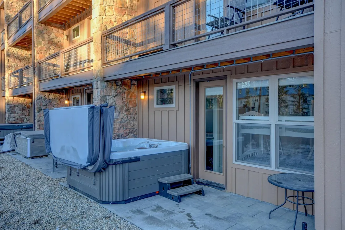 An exterior view of a tranquil patio area featuring a hot tub and a stone wall. The patio area looks spacious, ideal for al fresco dining or morning coffee while enjoying the fresh mountain air. The setup suggests a smooth transition from indoor dining to outdoor gatherings, highlighting the lifestyle benefits of the property.
