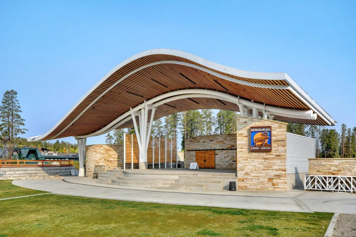 An energetic outdoor pavilion with a curved roof structure, stone façade, and wooden accents set in a community space. The area is designed for outdoor events, concerts, and communal gatherings, with open air seating for attendees. The mood is lively and welcoming, perfect for summer concerts or seasonal festivals.