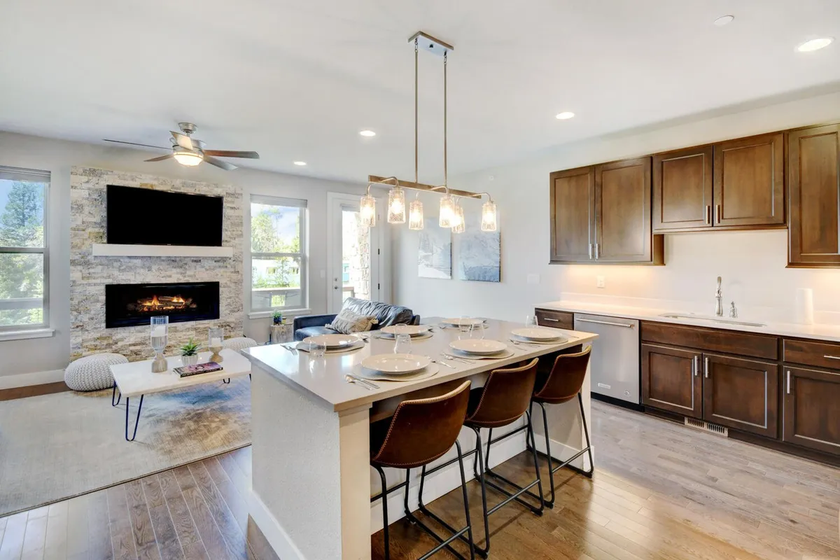 A spacious open-concept living area featuring a kitchen island with seating, a striking stone fireplace, and large windows flooding the space with natural light. High-quality furnishings invite relaxation, and the view through the windows connects guests to the mountain scenery outside.
