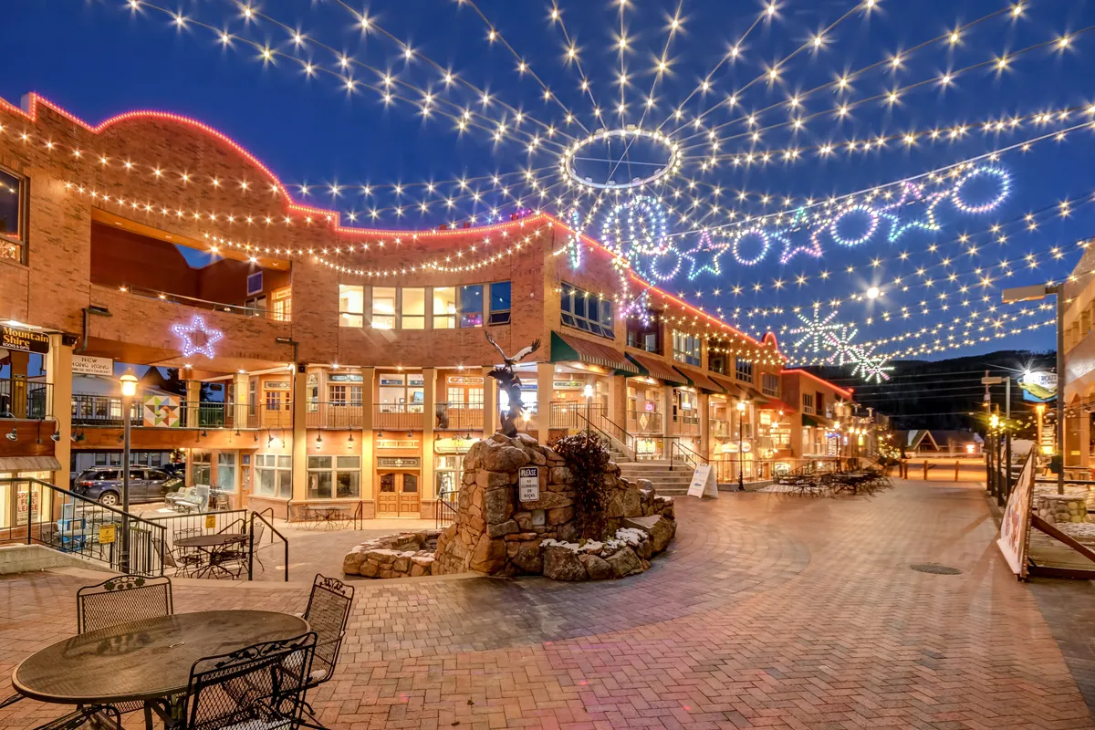 This image shows an outdoor plaza adorned with string lights and decorative stars, centered around a stone fountain. There are outdoor seating areas where people can dine, socialize, or enjoy events. The festive ambiance suggests a lively night market or seasonal celebration, with shops and restaurants lining the space.
